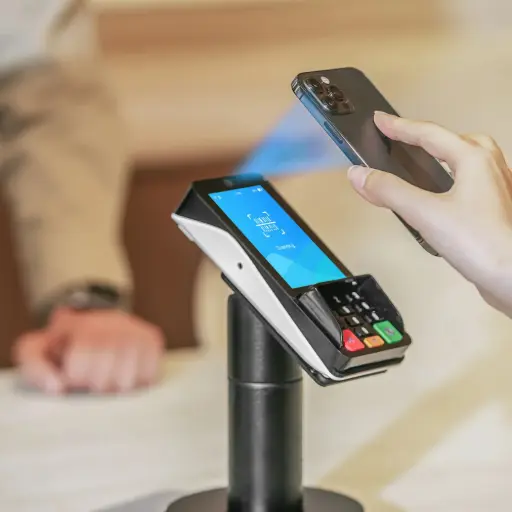 L'image montre une scène de paiement sans contact, où un client utilise un smartphone pour payer sur un terminal de paiement PAX A35 noir, qui est monté sur un support de comptoir. Le texte à droite, titré 'PIN Pad ou TPA ? La double fonctionnalité du PAX A35', explique la 'nature hybride' de cet appareil. Il est précisé qu'il peut à la fois servir de 'PIN Pad client' sécurisé (en 'mode semi-intégré', relié à une caisse qui envoie les montants) et de 'Terminal de Paiement Électronique (TPE)' moderne. Grâce à son 'OS PayDroid (basé sur Android)', il est également capable de fonctionner en 'mode TPE Autonome (TPA)', gérant ainsi l'intégralité du cycle de paiement (saisie du montant, transaction, télécollecte) de manière indépendante. Le texte conclut que cette 'double fonctionnalité' fait du PAX A35 une 'solution flexible et évolutive', s'adaptant à toutes les configurations de caisse.