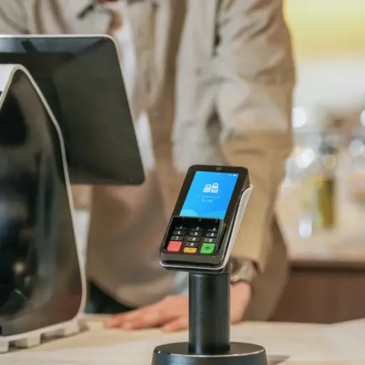 L'image présente le PIN Pad PAX A35 en situation d'utilisation dans un environnement commercial. Sur la gauche, le terminal est monté sur un support noir, posé sur un comptoir clair, avec l'écran affichant une icône de cadenas et un montant de "£0.00", suggérant qu'il est prêt pour une transaction. L'arrière-plan flou montre une personne (probablement un caissier ou un vendeur) et une partie d'un autre équipement de caisse, accentuant le contexte de point de vente. Le texte à droite de l'image, intitulé "PIN PAD PAX A35 : Pour quel secteur d'activité ?", explique que le PAX A35 est idéalement conçu pour le commerce de détail, en particulier dans les environnements à fort volume de transactions et ceux utilisant des systèmes de caisse intégrés. Le PAX A35 est retrouvé dans la grande distribution et les supermarchés, où la vitesse et la gestion des files d'attente sont cruciales. Il est également adapté à la restauration (restauration rapide, cafés), où son écran tactile permet de gérer facilement le pourboire, les programmes de fidélité et la lecture des codes QR. Dans le secteur de la mode et des boutiques spécialisées, son design moderne et la qualité de son écran de 4 pouces sont utilisés pour renforcer l'image de marque et afficher des promotions ciblées. Enfin, pour les pharmacies et services, il est polyvalent pour l'encaissement et peut s'intégrer à des solutions spécifiques. Le texte conclut que le A35 est la solution de choix pour tout professionnel souhaitant transformer son comptoir de caisse en un point de vente intelligent, capable d'accepter tous les modes de paiement modernes (cartes, mobiles, titres restaurant, Sans Contact Plus) de manière fluide et sécurisée.