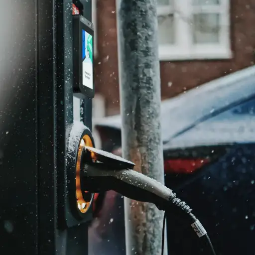 L'image montre une borne de recharge pour véhicule électrique dans un environnement extérieur enneigé, avec le pistolet de charge noir branché dans la prise lumineuse de la borne. Le texte à droite, titré 'Pourquoi choisir le PAX IM25 pour vos automates ?', s'adresse aux gérants de distributeurs automatiques, de 'bornes de recharge pour véhicules électriques' (comme celle de l'image), d''horodateurs', de 'stations de lavage automobile', de 'laveries automatiques' ou de 'consignes intelligentes'. Il est affirmé que le PAX IM25 est 'le terminal de paiement qu'il vous faut' pour ces points de vente libre-service. La section 'Ce qui fait la force de l'IM25' (surlignée en jaune) met en avant son 'écran tactile en verre trempé' ultra-résistant et son 'capteur de proximité intelligent' qui allume l'écran à l'approche du client. Le texte souligne que le PAX IM25 est 'conçu pour durer' et résister aux 'températures extrêmes' et à l''usage intensif', même 'en extérieur ou en environnement hivernal'. Une note finale 'Bon à savoir' précise que si les clients ont besoin d'insérer leur carte, il faut privilégier le PAX IM30, qui est équipé d'un lecteur de carte à puce et bande magnétique.